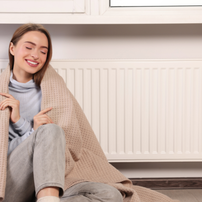 A young woman sits on the floor, wrapped in a blanket, near a wall radiator unit