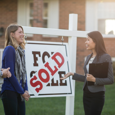 A couple stand in front of a fore sale sign with a 'sold' sign pasted into it, speaking with a realtor