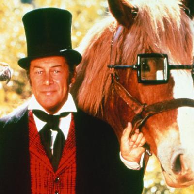 Actor Rex Harrison wearing a long black coat, a red waistcoat, and a black top hat, with a parrot sitting on his right hand as he stands beside a horse wearing a pair of glasses in a publicity portrait for the 1967 film 'Doctor Dolittle.'