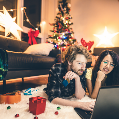 A couple lie on the carpet in their sitting room and shop online from a laptop; a Christmas tree is lit in the background