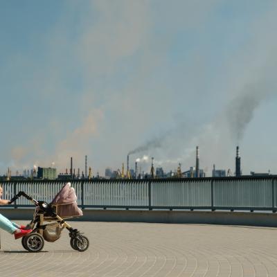 Parent with baby sitting on a bench as air behind them is polluted by smoke stacks.