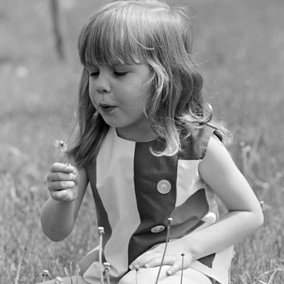 Child in the '60s blows on a dandelion while sitting in the grass.