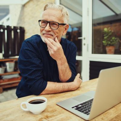 Retired person sits in their backyard drinking coffee in front of their laptop.