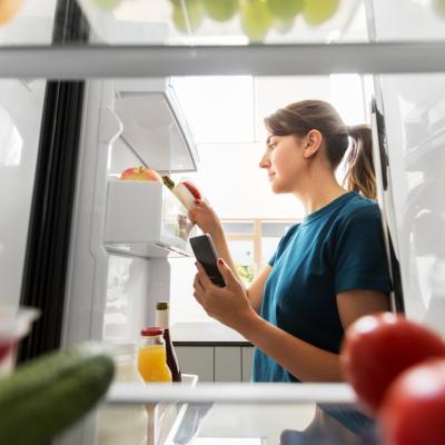 Person looking at the food in the door of their fridge with a smartphone in their hand.