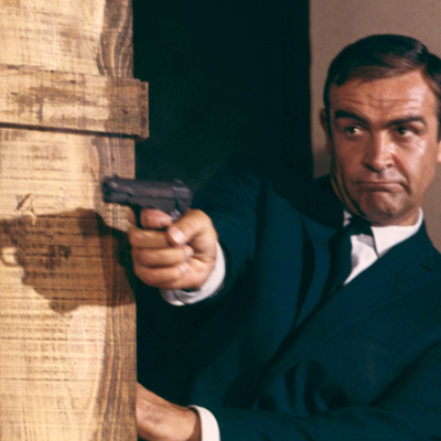 Sean Connery as James Bond 007 firing a pistol behind crates from the film "Goldfinger", 1964.