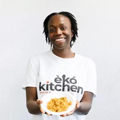 Simileoluwa Adebajo wears a shirt from her business Eko Kitchen and holds a plate of food, standing in front of white background.