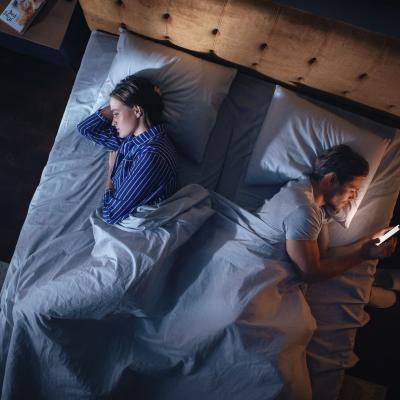 Overhead view of couple laying in bed facing away from each other in the dark while one person is scrolling on phone.