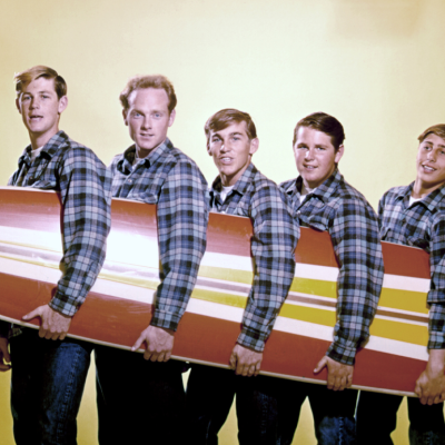 Rock and roll band "The Beach Boys" pose for a portrait with a surfboard in August 1962 in Los Angeles, California. (L-R) Brian Wilson, Mike Love, Dennis Wilson, Carl Wilson, David Marks. 