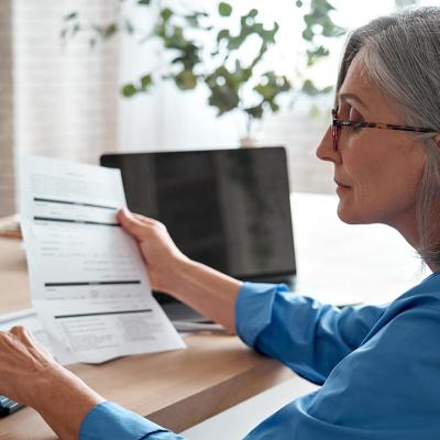 A middle-aged woman sitting with a laptop and holding a bill while entering numbers in a calculator.