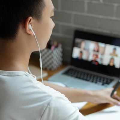 Young person with headphones works on computer with virtual meeting, in dorm setting.