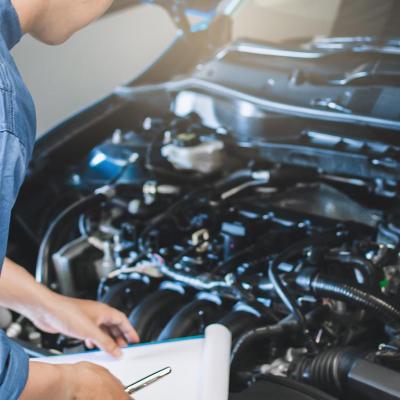 Mechanic in blue suit with clipboard inspects under the hood of car.