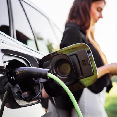 A woman with a phone is leaning on her electric car while it is being charged.