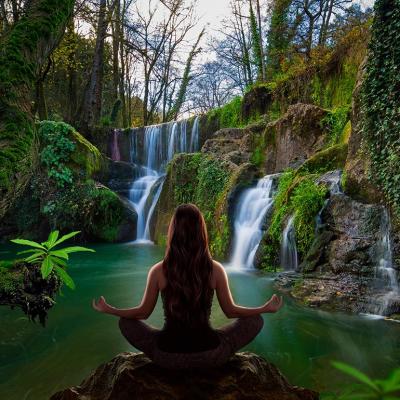 Person sitting in yoga lotus pose facing away from viewer looking at waterfall. 