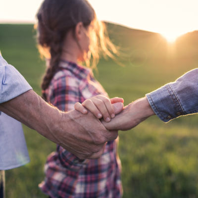 A small girl with her senior grandparents holding hands outside in nature at sunset.