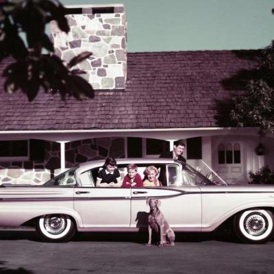 A family of four in their Mercury Monterey automobile in the driveway of their home with their pet dog sitting next to the car.