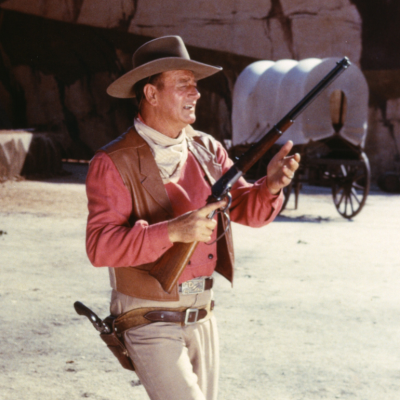 John Wayne (1907-1979), US actor, wearing a tan leather waistcoat over a pink shirt, taking aim with a rifle, in a publicity still issued for the film, 'Rio Bravo', USA, 1959. 