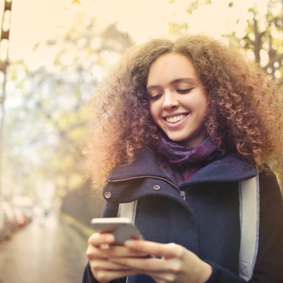 A smiling young woman looks at her smartphone while outside.