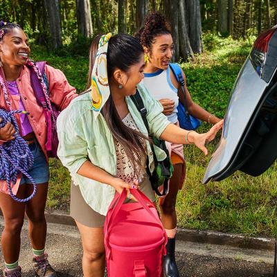 Three young people carrying bags for a summer trip load a car trunk.