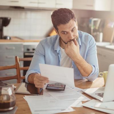 A man sitting in a kitchen with a laptop and calculator on the table while reading a letter with a focused gesture.