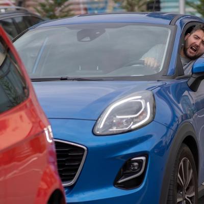 A man driving inside a blue car, his head is sticking out and is seen yelling at a red car in front of him.