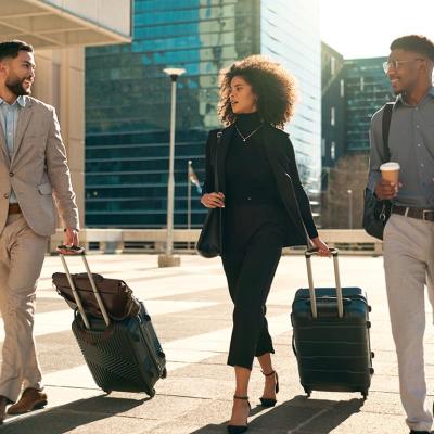 Three professionals in business attire are walking with their suitcases while seemingly having a chat.