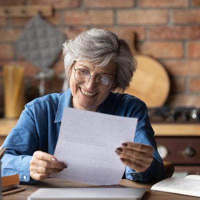 An elderly lady is sitting in her kitchen while happily reading a piece of paper.