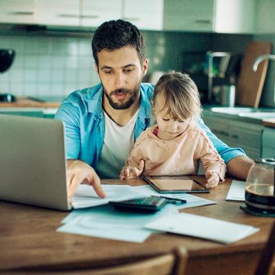 Father sitting at kitchen table with bills, calculator and laptop computer while holding small child.