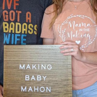 Self portrait of Kirsti and Justin Mahon from the neck down, holding a sign that says "Making Baby Mahon."