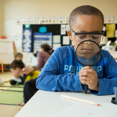 A child in a classroom looks through a magnifying glass down at learning materials on his desk. The magnifying glass makes the bottom of his face appear larger.