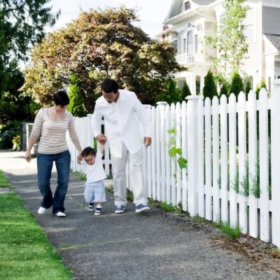 Family of three walking on the sidewalk surrounded by a white picket fence.