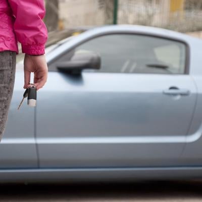 Person in dark pink shirt and black jeans holding car keys seen from behind, standing in front of used light blue car. 