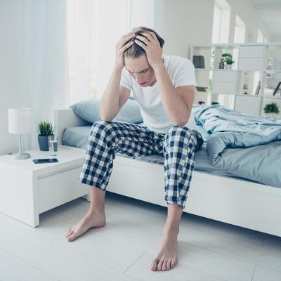 Young adult male sitting on a bed with both hands holding his head seemingly having a headache.