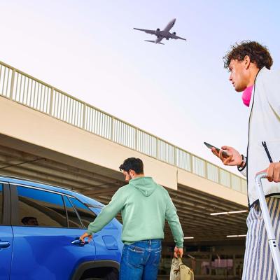 Two travelers head to a blue car with luggage while a plane flies over head.