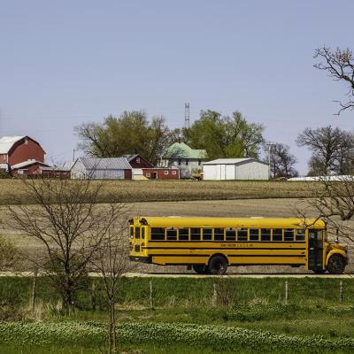 KINGSTON, ILLINOIS A public school bus passes by a large farmstead along a rural road on a spring day, with the edge of a county forest preserve in the foreground.