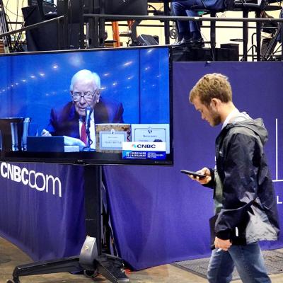 Warren Buffett appears on a TV screen at the Berkshire Hathaway sharesholders meeting, while a bystander walks past.