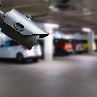 Close up of a security camera in focus inside an indoor parking garage, with blurred cars in the background.