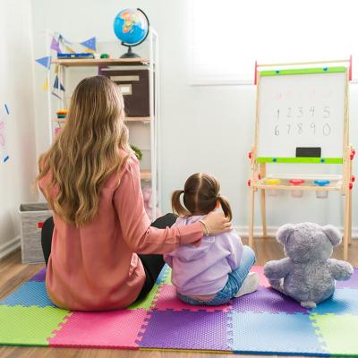 Preschooler in classroom with adult figure sitting on mat as seen from the back, next to a plush toy. 