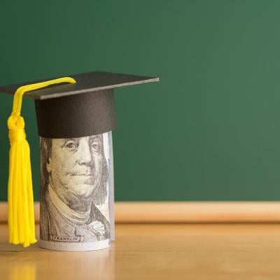 Concept of student debt illustrated by a rolled up dollar bill wears graduation cap with a chalkboard background in classroom.