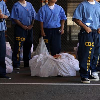 Inmates at San Quentin State Prison shown from the neck down wearing blue uniforms and navy sweat pants that say "CDCR prisoner" wait in line in 2016.