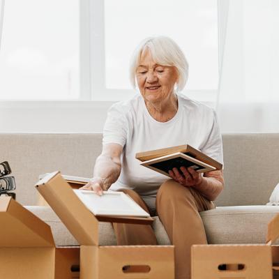 An elderly woman sitting on a sofa collecting home items to put inside boxes.