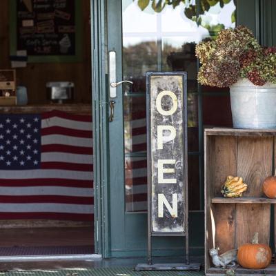 An "open" sign hangs in a shop window with fall decor and American flag in the background.