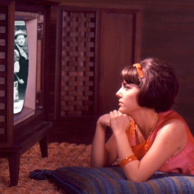 A composite image shows a young person in a pink and orange dress watches an episode of 'The Honeymooners' on a console TV set in the late 1960s or early 1970s.