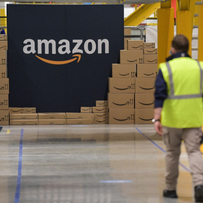 An Amazon warehouse employee walking towards a pile of Amazon-branded boxes and a large Amazon logo sign