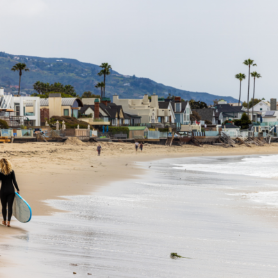 A surfer in a wet suit walking along the beach in Malibu, California carrying a surfboard.