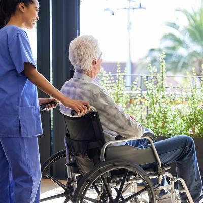 A senior man in a wheelchair looking out a balcony and assisted by a nurse.