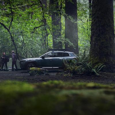 Dark gray Mazda car parked in a forest where two adults are setting up a tent.