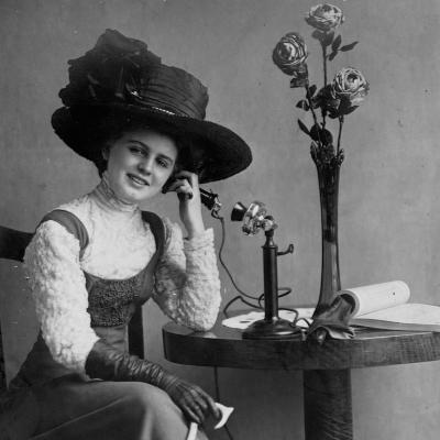 Person with hat seated at table talking on an early standing telephone.