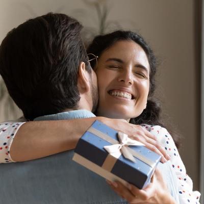 Woman smiling with eyes closed hugging a man whose back is to the camera, she is holding a small blue box tied with a bow.