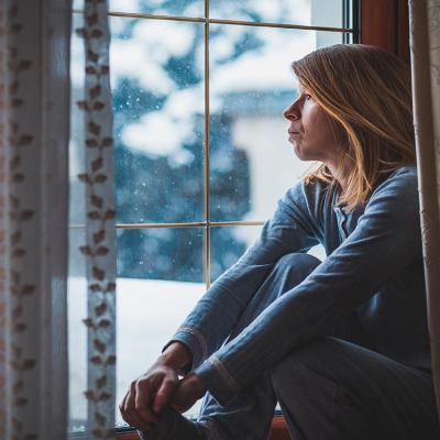 A woman sadly sitting by and looking out a window during winter.