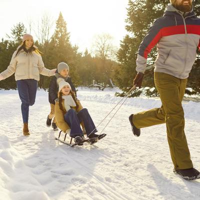 Two parents running outdoors and their two children in sleds during wintertime.